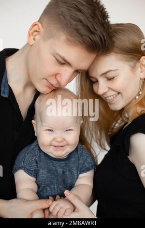 Ritratto di famiglia felice da tre in nero guarda su sfondo bianco. Mamma e papà toccano delicatamente le mani piccole di gioioso sorridente bambino primo piano Foto Stock