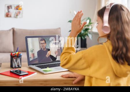 La bambina della scuola sta studiando a distanza, usando la webcam del laptop e le cuffie, alza la mano per rispondere alla domanda dell'insegnante. Scuola domestica e concetto di formazione a distanza. Tutor online che ha lezioni a distanza Foto Stock