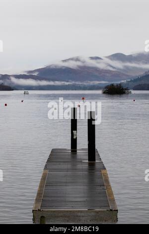 Molo sul lago Windermere con nebbia che si avvolse sulle montagne in una giornata nuvolosa Foto Stock