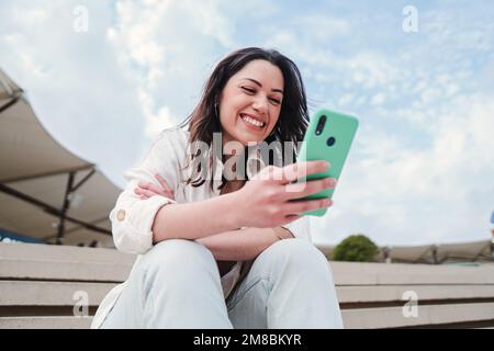Primo piano ritratto di giovane donna brunetta che guarda i contenuti su un cellulare app seduto all'aperto. Donna spensierata sorridente e divertente SMS messaggi con uno smartphone. Foto di alta qualità Foto Stock