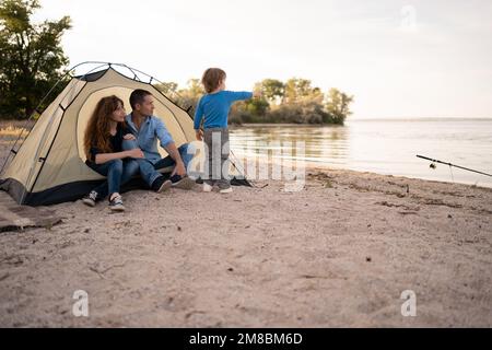 famiglia felice nella tenda al campeggio che punta il dito a qualcosa. camping, turismo, escursione e concetto di persone Foto Stock