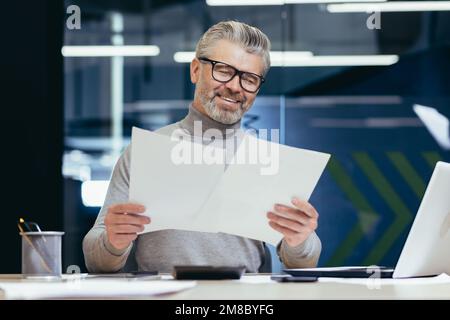 Uomo d'affari senior sorridente dai capelli grigi che lavora in ufficio alla scrivania con computer portatile e documenti. Tiene in mano, guarda fogli di carta, accordi, fatture. Foto Stock