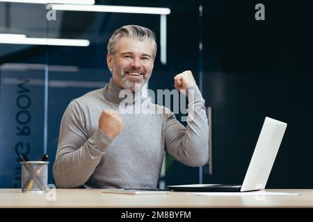 Ritratto di un vecchio uomo di successo dai capelli grigi. Un uomo felice è seduto in ufficio al tavolo con un computer portatile. Celebra la vittoria, il successo, sorride alla telecamera, mostra forza con le mani. Foto Stock