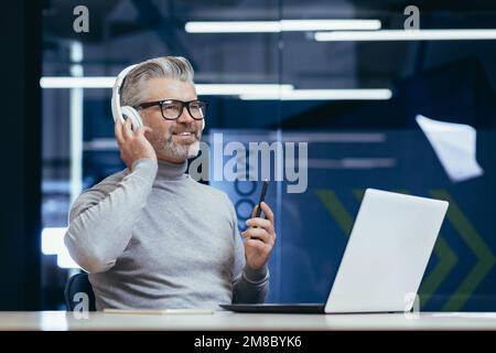 Uomo anziano sorridente dai capelli grigi seduto alla scrivania in ufficio con cuffie davanti al computer portatile. Parla tramite videochiamata, ascolta musica, webinar, studi, insegna, riposi. Foto Stock