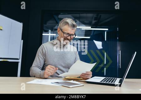 Uomo anziano dai capelli grigi che lavora in ufficio alla scrivania con computer portatile e documenti. una busta con una lettera, fatture e rapporti, buone notizie. Foto Stock