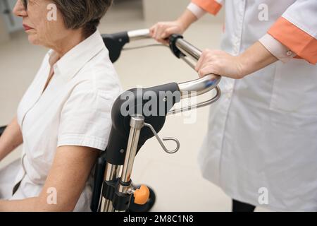 La donna anziana viene trasportata in sedia a rotelle da un operatore sanitario Foto Stock