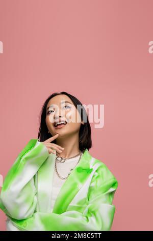 donna asiatica sognante e sorridente in giacca alla moda toccando mento e guardando isolato su rosa, immagine stock Foto Stock