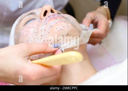 Un estetista rimuove una maschera idratante del foglio sul viso di una donna con una spatola, procedure cosmetologiche. Foto Stock