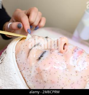 Un estetista rimuove una maschera idratante del foglio sul viso di una donna con una spatola, procedure cosmetologiche. Foto Stock