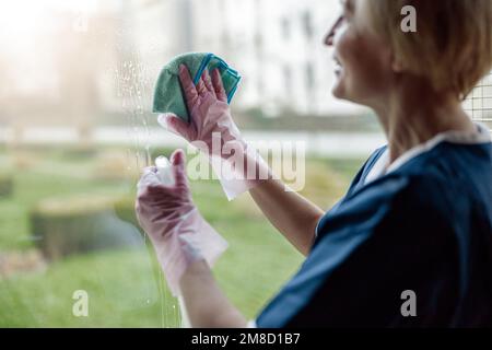 Cameriera senior in guanti e finestra di pulizia uniforme con panno e detergente spray Foto Stock