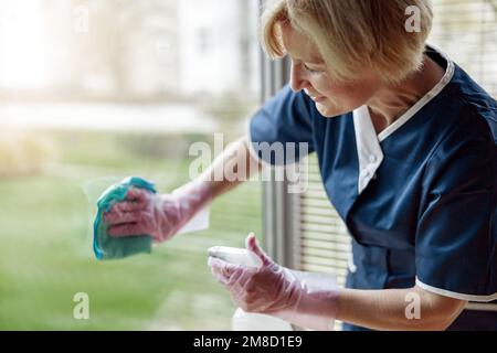 Cameriera senior in guanti e finestra di pulizia uniforme con panno e detergente spray Foto Stock