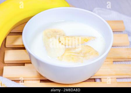 Fette di banana in latte di cocco in una ciotola bianca su sfondo di legno chiaro. Foto Stock