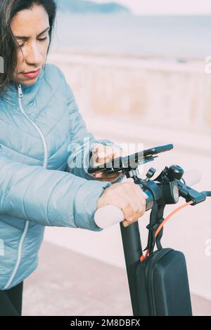 Vista laterale della bella donna latina in sella al suo scooter motorizzato in città, attivazione di app per smartphone, concetto di mobilità trasporto sostenibile, zero Foto Stock