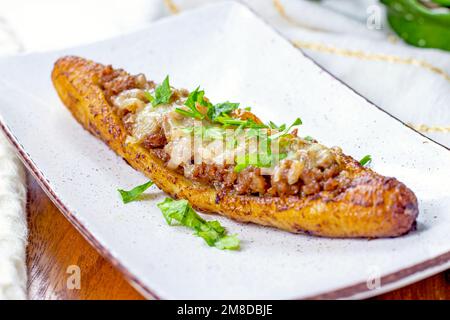 Canoe di piantana mature dolci al forno (Canoas de Platanos Maduros) farcite con carne macinata, formaggio e verdure su un piatto su sfondo di legno. Foto Stock