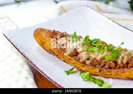 Canoe di piantana mature dolci al forno (Canoas de Platanos Maduros) farcite con carne macinata, formaggio e verdure su un piatto su sfondo di legno. Foto Stock