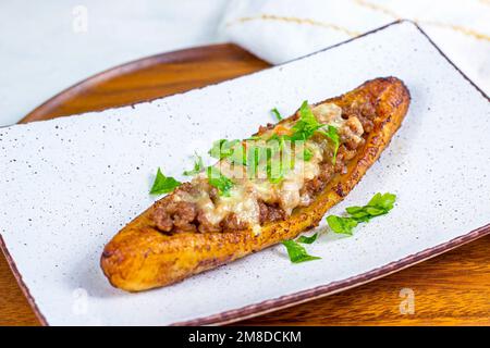 Canoe di piantana mature dolci al forno (Canoas de Platanos Maduros) farcite con carne macinata, formaggio e verdure su un piatto su sfondo di legno. Foto Stock