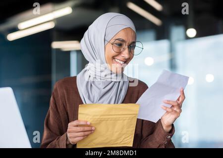 Donna d'affari in hijab primo piano sulla notifica di lettura, donna musulmana ha ricevuto la lettera della busta postale con il messaggio che legge gioiosamente e sorridente, donna riuscita al lavoro all'interno dell'ufficio. Foto Stock