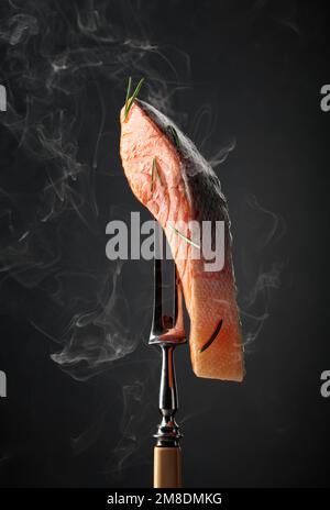 Salmone affumicato con rosmarino su forchetta. Pezzo di pesce con fumo naturale su sfondo nero. Foto Stock