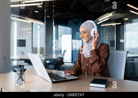 Donna d'affari moderna di successo che lavora all'interno dell'ufficio con un computer portatile, una donna musulmana che indossa un hijab e cuffie che ascolta audiolibri e podcast sul posto di lavoro, donna soddisfatta e di successo. Foto Stock
