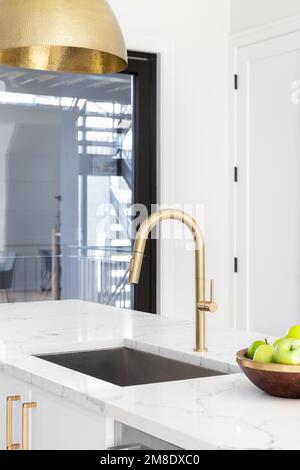 Un rubinetto d'oro da cucina in una cucina bianca con un piano di marmo, lampada d'oro appesa giù, e una ciotola di mele verdi. Foto Stock