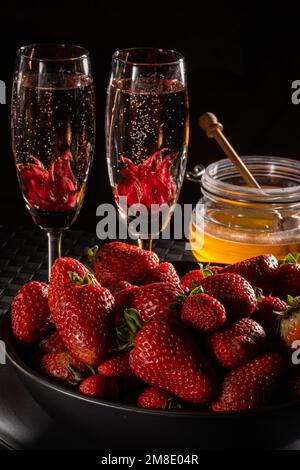 Una romantica scena di champagne, fiori di ibisco, fragole e miele ambientata in una scena buia. Ideale per menu, siti Web o pubblicazioni. Foto Stock