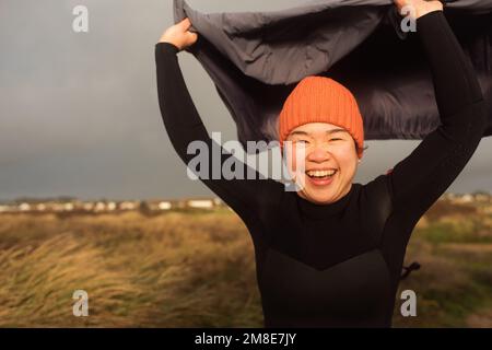 Donna asiatica che tiene il suo cappotto soffiato nel vento alla spiaggia Foto Stock