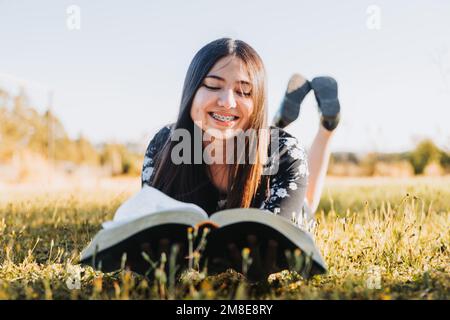 Sorridente ragazza cristiana sdraiata sull'erba che ha comunione con dio leggendo la sua bibbia, nel campo al tramonto. Foto Stock