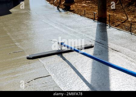 Il lavoratore tiene la cazzuola in acciaio fresno e leviga il marciapiede appena versato su calcestruzzo bagnato in cui la costruzione sta avvenendo Foto Stock