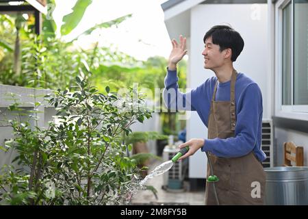 Sorridente maschio asiatico in grembiule innaffiando le piante al cortile e salutando il suo vicino con il volto sorridente amichevole Foto Stock