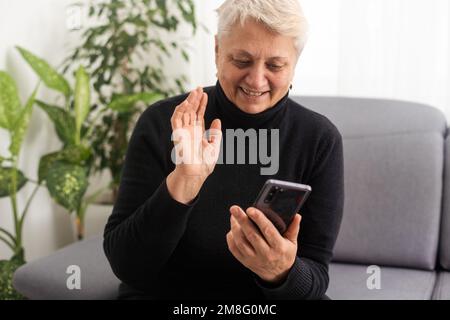 Felice donna sorridente più anziana utilizzando il telefono, guardando lo schermo, seduto su un comodo divano in soggiorno, entusiasta senior femmina chat in rete sociale Foto Stock