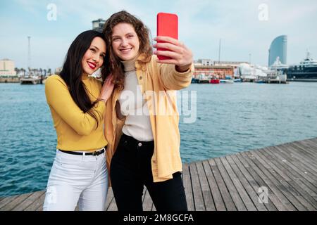 Le giovani donne sorridono mentre scattano selfie con un telefono cellulare all'aperto sul lungomare. Foto Stock