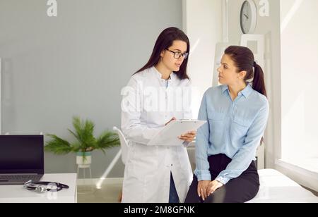 Giovane donna medico in cappotto medico bianco consulta paziente femminile siede sul divano dell'ospedale Foto Stock