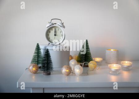 Una sveglia bianca, un puntatore a dodici, un piccolo albero di Natale artificiale verde e candele accese in candelabri su una cassettiera bianca. Copia Foto Stock