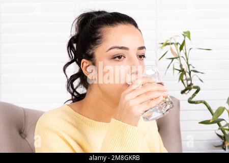 Ritratto di bruna donna che beve acqua. La ragazza caucasica gode di acqua minerale pura e fresca. Poltrona. Signora assetata che tiene un bicchiere di acqua ancora. Foto Stock