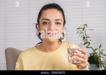 Donna brunetta sorridente che tiene un bicchiere d'acqua. Felice ragazza caucasica millennial guardando macchina fotografica. Acqua minerale pura fresca, concetto di cura del corpo e della pelle. Foto Stock