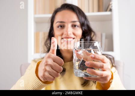 Donna che tiene un bicchiere d'acqua. Sorridente ragazza caucasica felice consiglia di bere ancora acqua minerale mostrando pollici in su. Concetto di stile di vita sano. Foto Stock