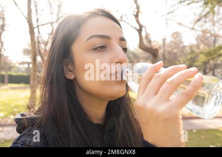 Primo piano immagine scattata dalla testa di una donna che beve acqua. Donna che tiene una bottiglia di vetro in piedi all'aperto. La ragazza beve ancora acqua pulita per la dieta, la cura della pelle Foto Stock