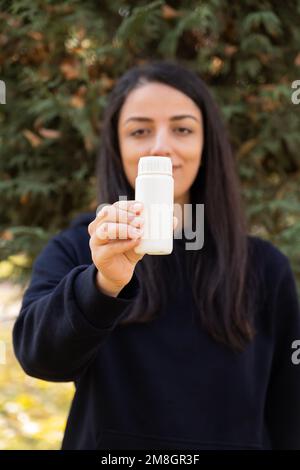 Mostrando la bottiglia della pillola. Immagine verticale della ragazza caucasica in piedi fuori, bianco vuoto vaso di plastica mock su sulla sua mano. Consigliare la medicina. Foto Stock