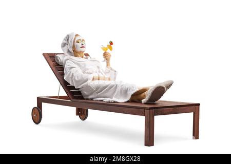 Donna in un accappatoio posata con una maschera viso e cetrioli sugli occhi e tenendo un bicchiere di cocktail isolato su sfondo bianco Foto Stock