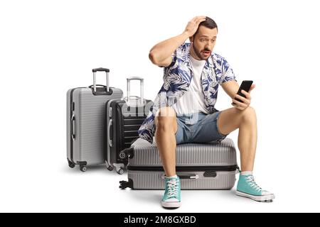 Scioccato turista maschile seduto su una valigia e guardando uno smartphone isolato su sfondo bianco Foto Stock