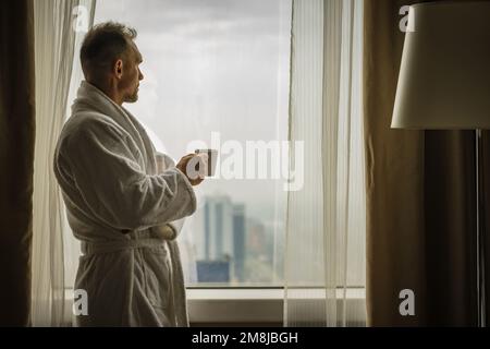 L'uomo caucasico si prepara per un nuovo giorno mentre è in viaggio d'affari. Guardando attraverso la finestra della camera dell'hotel e bevendo caffè Foto Stock
