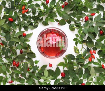 Tazza di tè di rosa rossa sullo sfondo con foglie fresche e bacche, vista dal tavolo. Tazza di vetro di tisana sana con rosee, foglie e fiori. Foto Stock