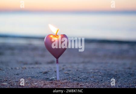 Candela a forma di cuore che brucia sulla sabbia della spiaggia vicino alle onde del mare durante l'alba del tramonto. Candela che brucia sotto forma di cuore in sabbia sullo sfondo del sole che sorge. Atmosfera romantica. St San Valentino. Foto Stock