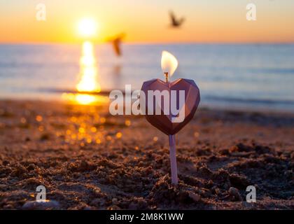 Candela a forma di cuore che brucia sulla sabbia della spiaggia vicino alle onde del mare durante l'alba del tramonto. Candela che brucia sotto forma di cuore in sabbia sullo sfondo del sole che sorge. Atmosfera romantica. St San Valentino. Foto Stock