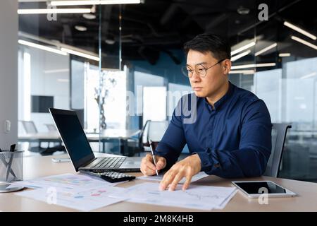 Concentrato giovane uomo d'affari asiatico lavora in ufficio al tavolo con computer portatile, scrive documenti, grafici, accordo, fatture. Foto Stock