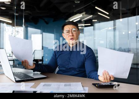 Problemi sul lavoro. Preoccupato giovane asiatico, uomo d'affari, freelance. Si siede in ufficio a un tavolo con un computer portatile e tiene i documenti in mano. Distende le mani, guarda la telecamera confusa. Foto Stock