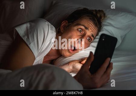 Ragazza sorpresa utilizzando lo smartphone a letto. Insonnia e concetto di dipendenza dai social media. Foto Stock