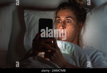 Donna scioccata che usa lo smartphone a letto. Insonnia e immagine concettuale stalking Foto Stock