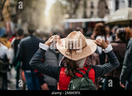 vista posteriore di una donna con cappello e zaino che cammina attraverso un mercato tradizionale in spagna. tiene il cappello. concetto di turismo urbano e vacanze. Foto Stock