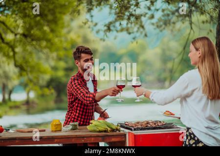 Felice coppia brindando bicchiere di vino rosso mentre si ha picnic francese cena festa all'aperto durante le vacanze estive vicino al fiume in bella natura Foto Stock